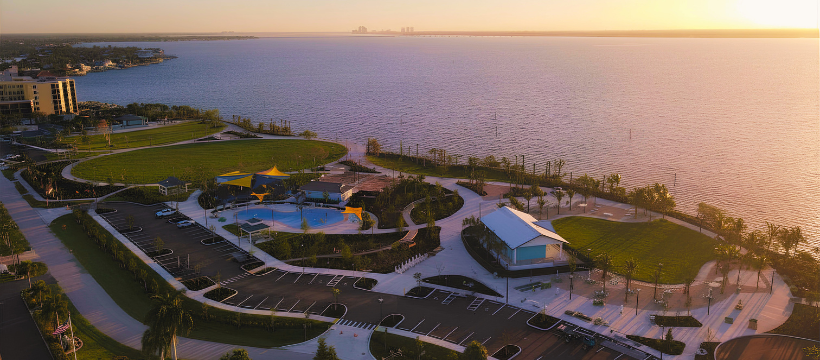 Sunrise aerial of Jaycee Park showing parking areas, lawns, splash pad, and riverfront promenade glowing in warm light.